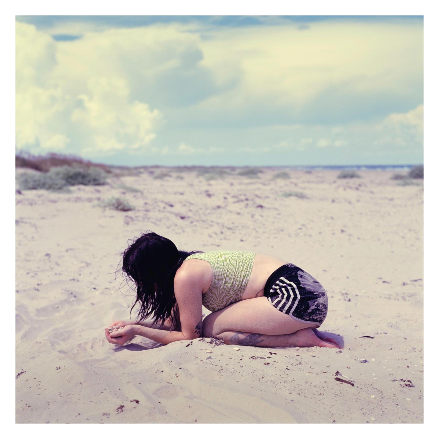 A woman kneels on a sandy beach, looking down at her hands.