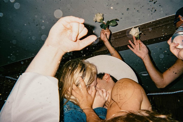 A couple kisses while surrounded by other people holding roses and a drink.