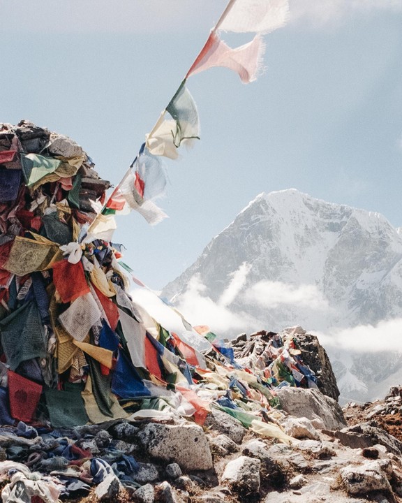 Colorful prayer flags are strung across a rocky mountain landscape with a snow-capped peak in the background.