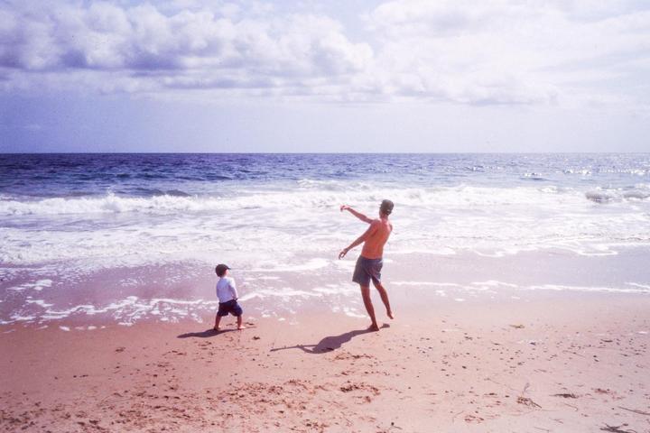 A man and a child stand on a sandy beach with waves crashing in the background under a cloudy sky.