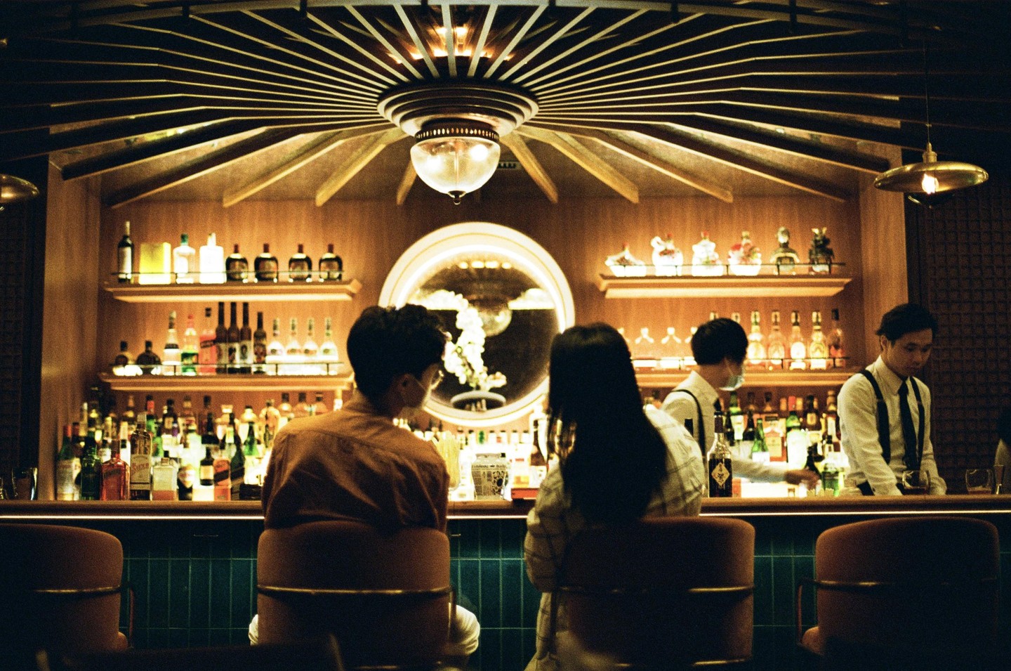 A couple sits at a bar with two bartenders behind the counter, under a decorative ceiling light.