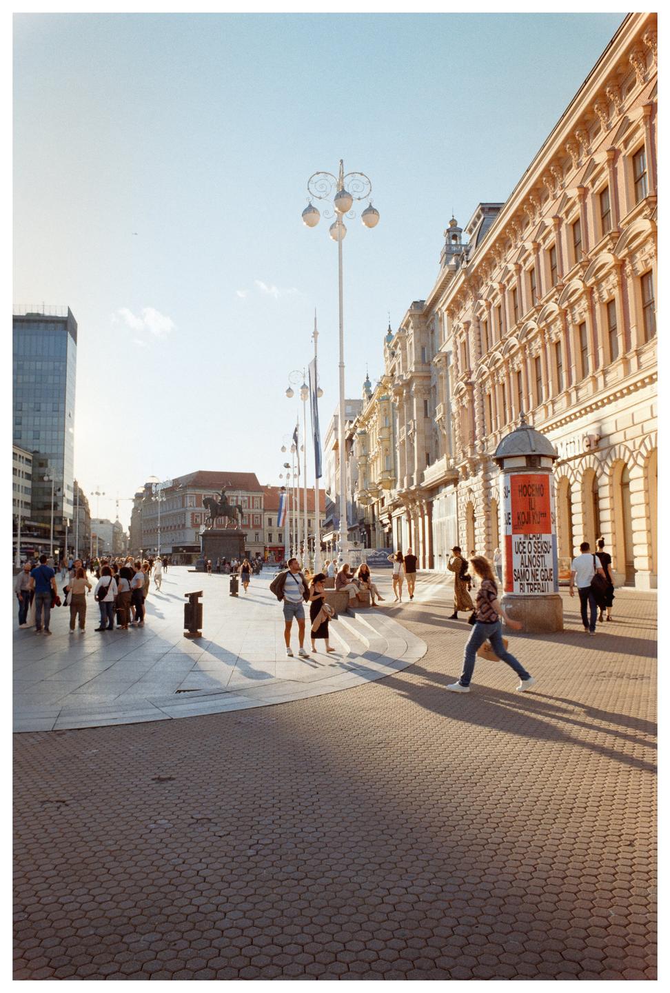 A wide street in a city is filled with people walking and standing, with buildings and a statue visible in the background.