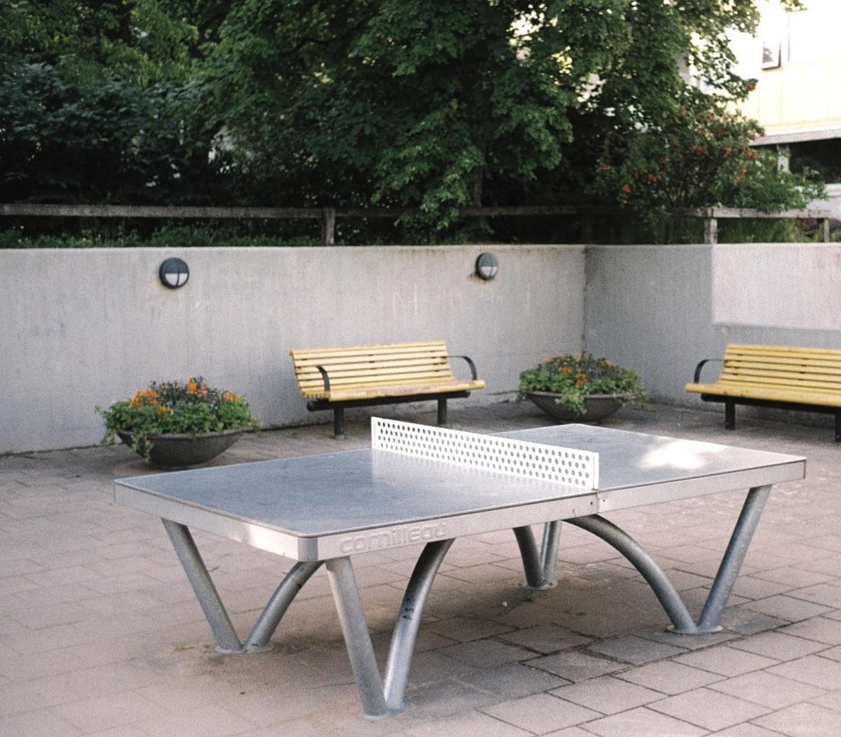 A ping pong table stands on a paved patio with two yellow benches and planters against a concrete wall.