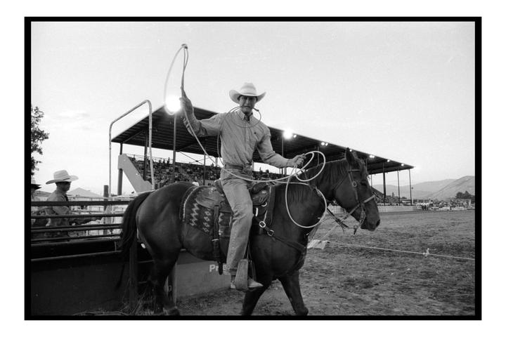 A cowboy on horseback holds a lasso in a rodeo arena with spectators in the background.