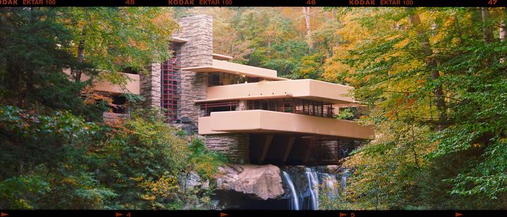 A modern house with multiple levels and balconies is built into a rocky cliff over a waterfall, surrounded by trees with green and autumn foliage.