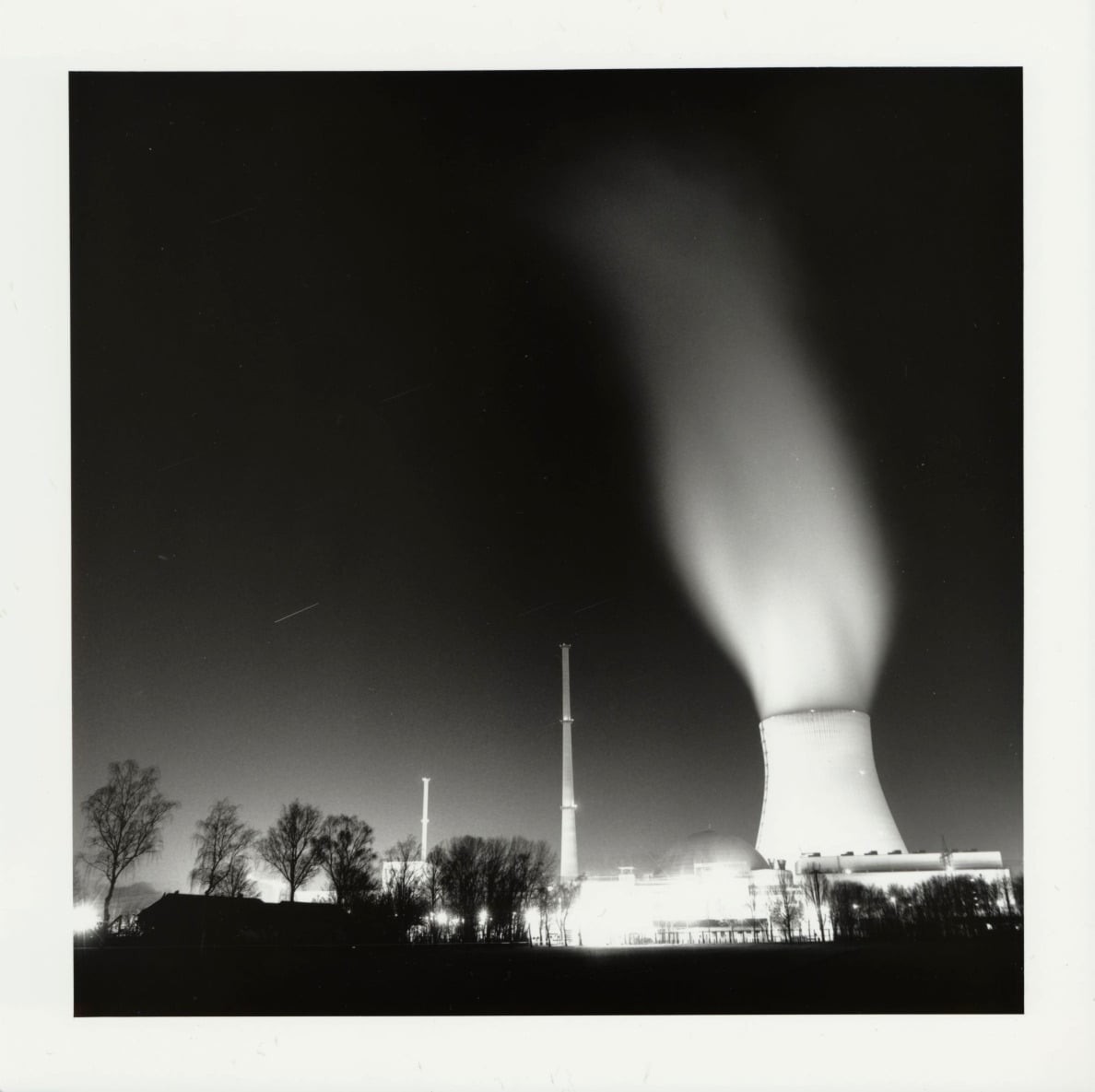 A nuclear power plant with a large cooling tower emitting steam is illuminated at night.