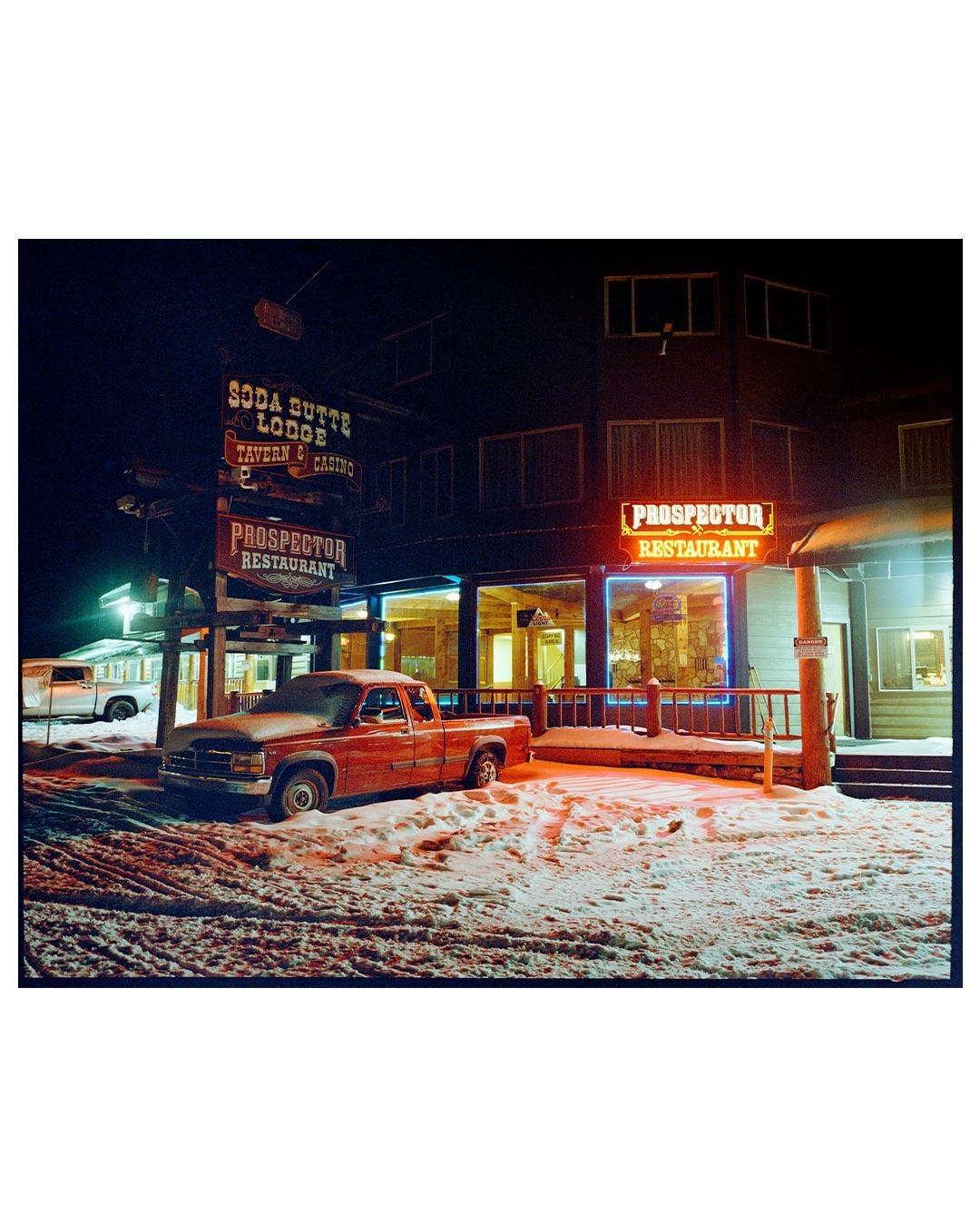 A red pickup truck is parked in the snow outside a lodge and restaurant at night.