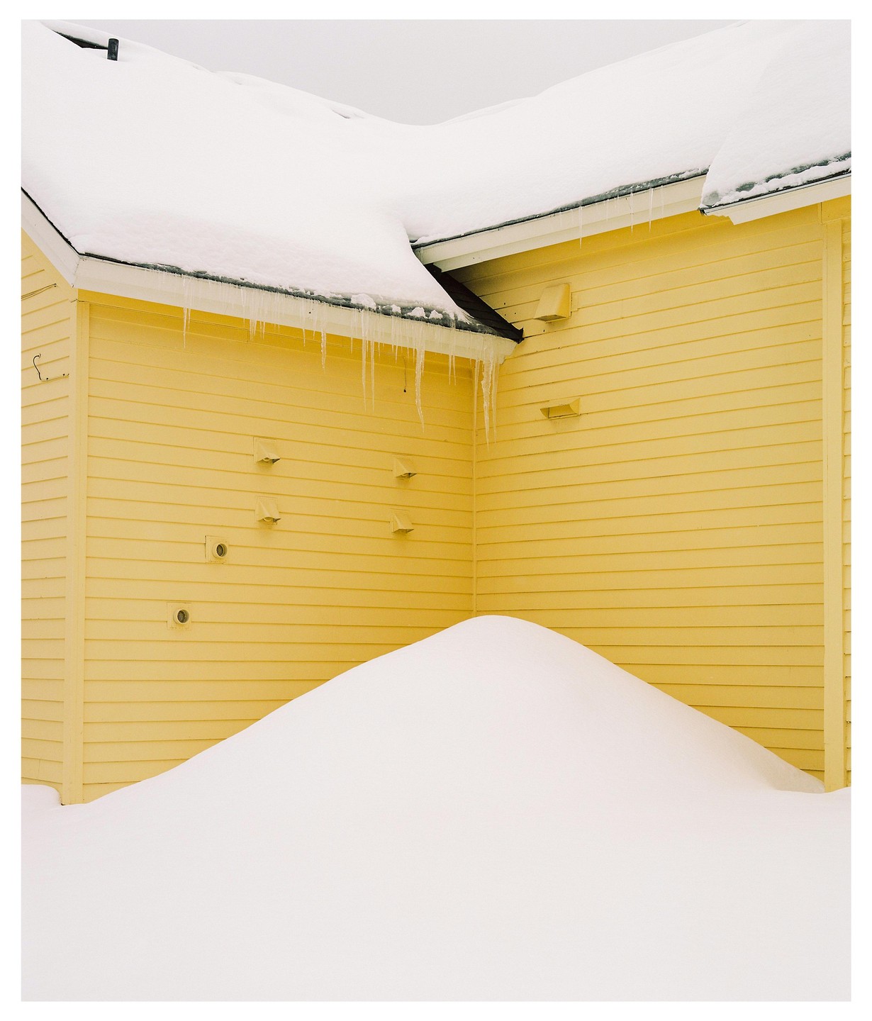 A large snowdrift is piled against the yellow side of a building with icicles hanging from the roof.