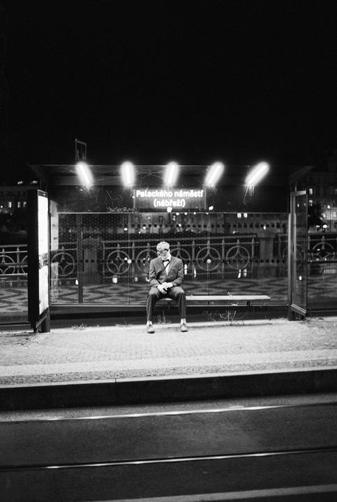 A man in a suit and bow tie sits on a bench at a bus stop at night.