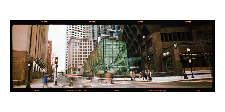 A street scene shows a modern glass building and other city buildings with blurred people and vehicles moving along the street.