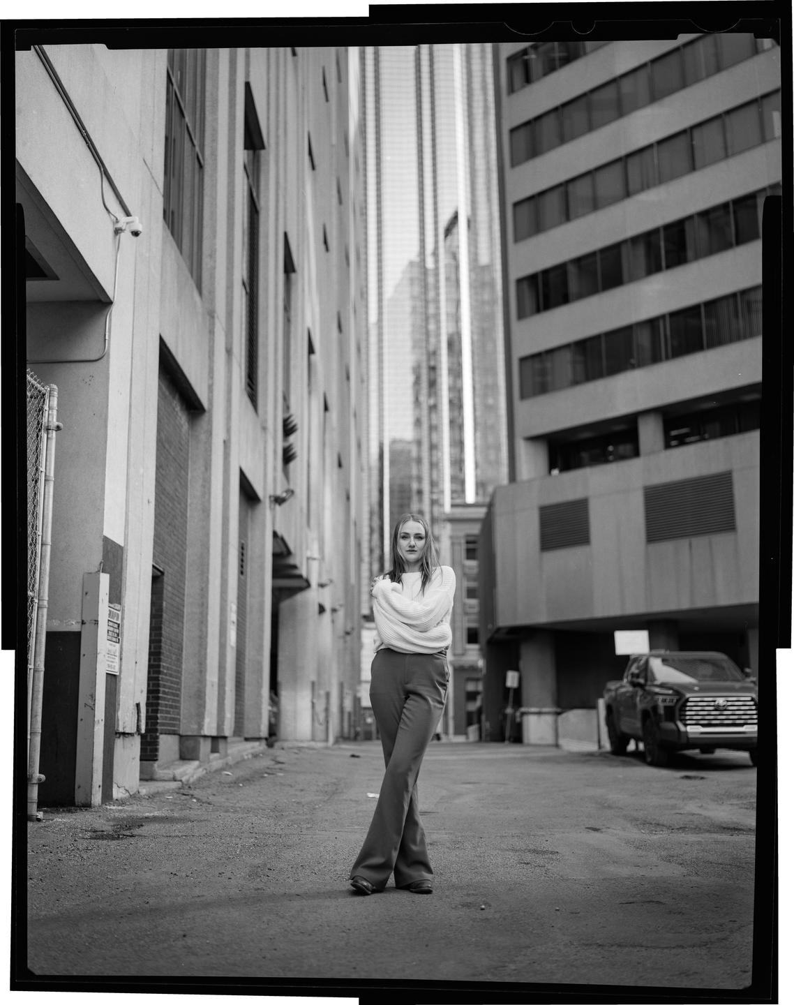A woman stands in an alley with her arms crossed, looking at the viewer.
