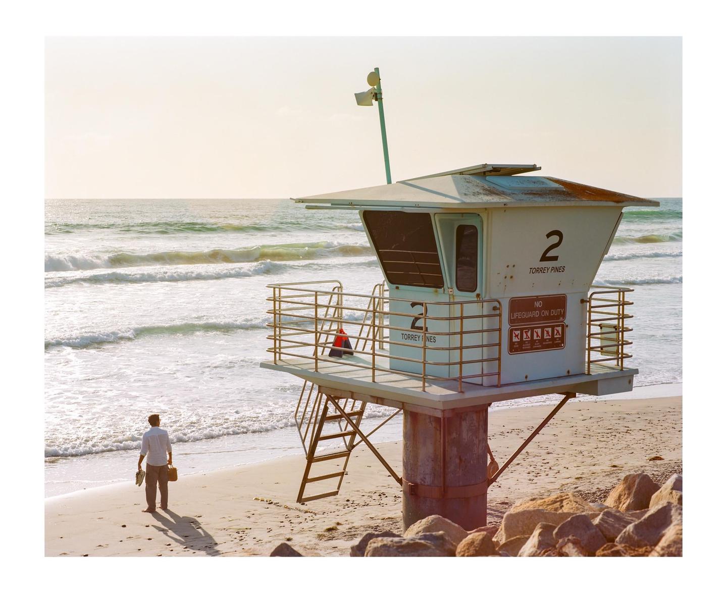 A man stands on a sandy beach next to a lifeguard tower as waves crash in the background.