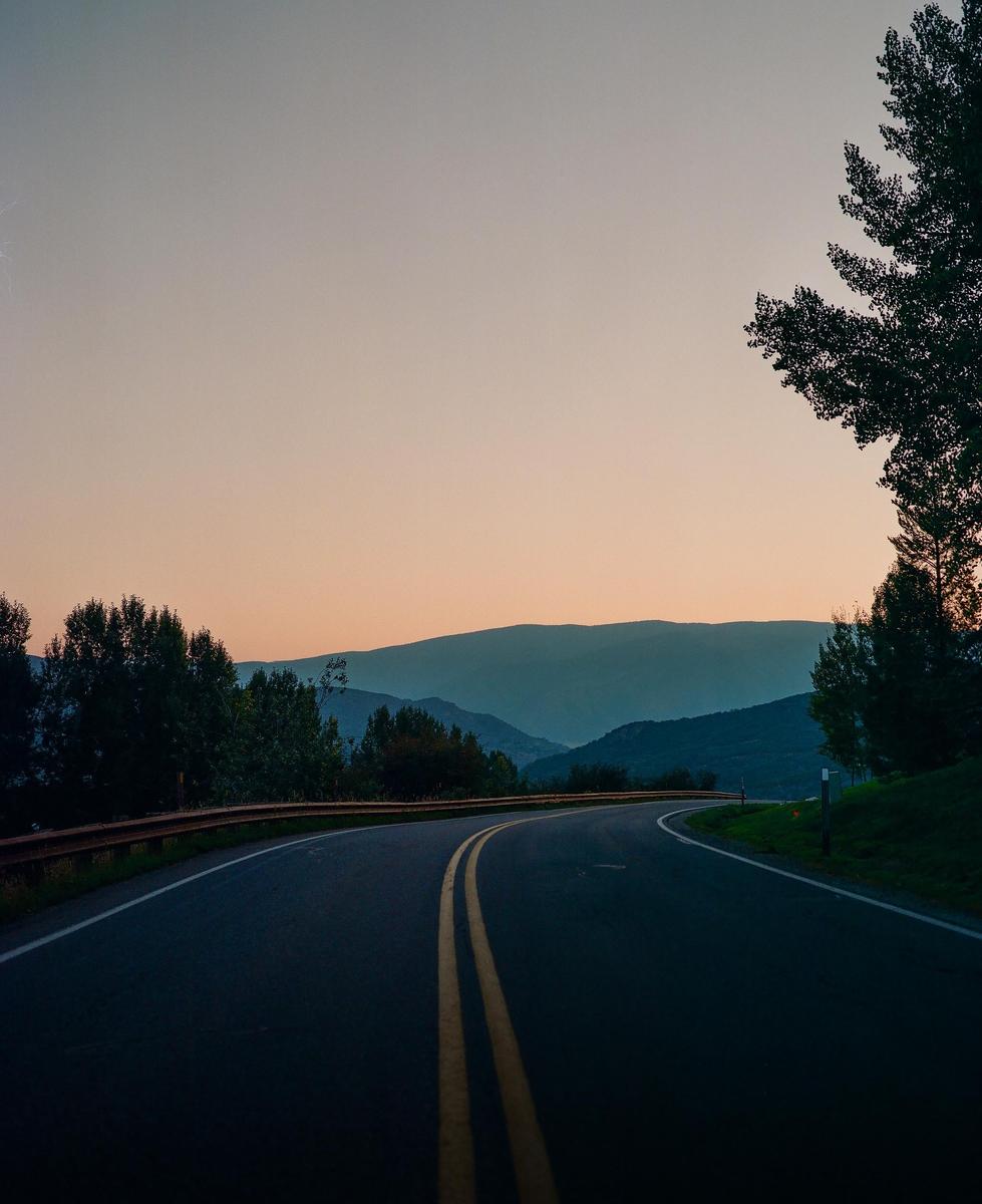 A winding road with a double yellow line leads into a mountain range at dusk.