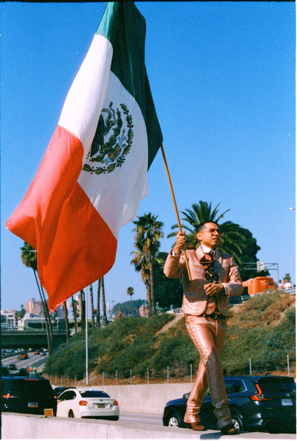 A man in a shiny suit holds a large Mexican flag on the side of a highway.