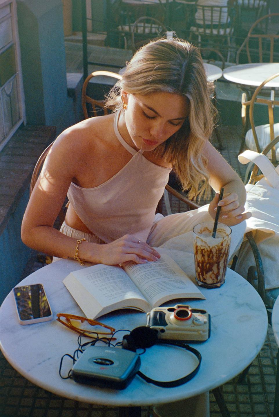 A woman reads a book at an outdoor cafe table with a drink, a phone, sunglasses, a camera, and a cassette player.