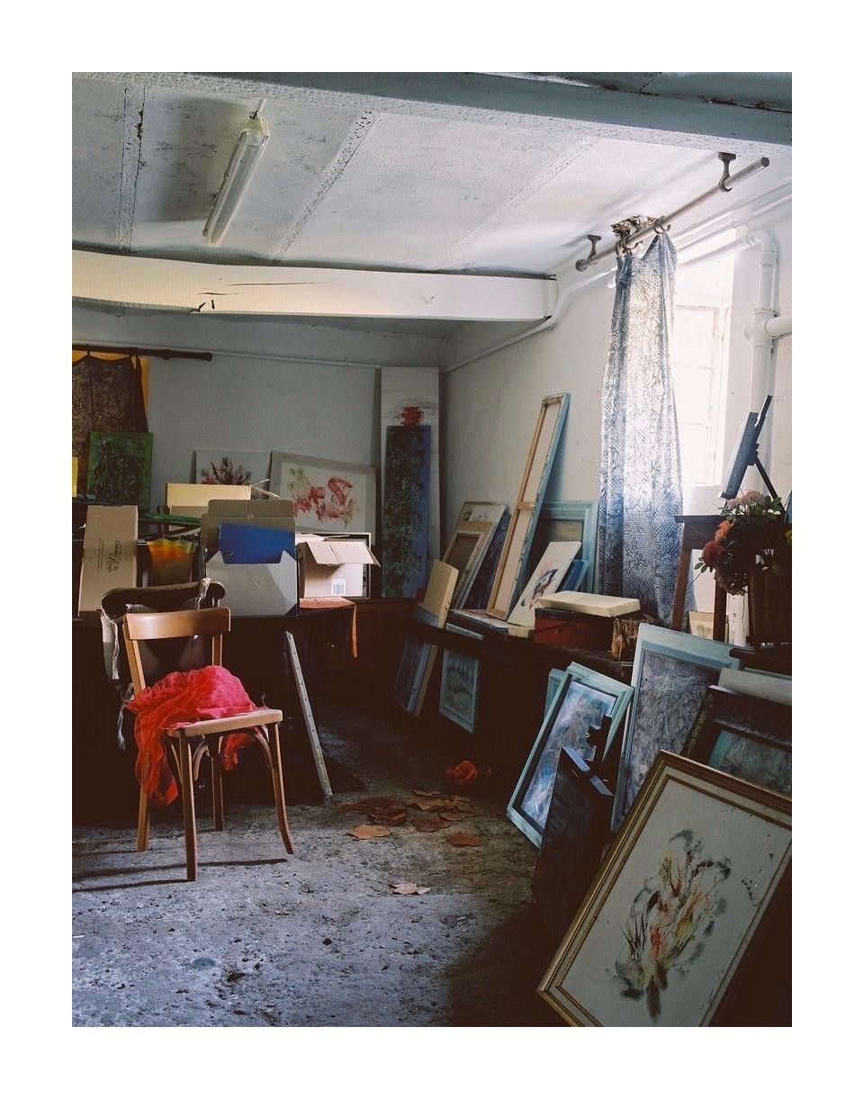 A wooden chair with a red cloth on it sits in a cluttered room filled with paintings, boxes, and other items.