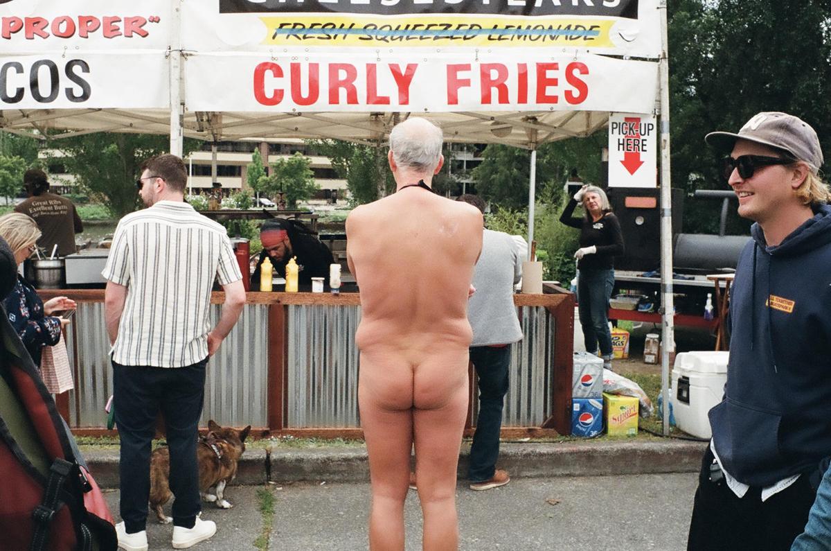 A nude man stands in front of a food stall with other people around him.