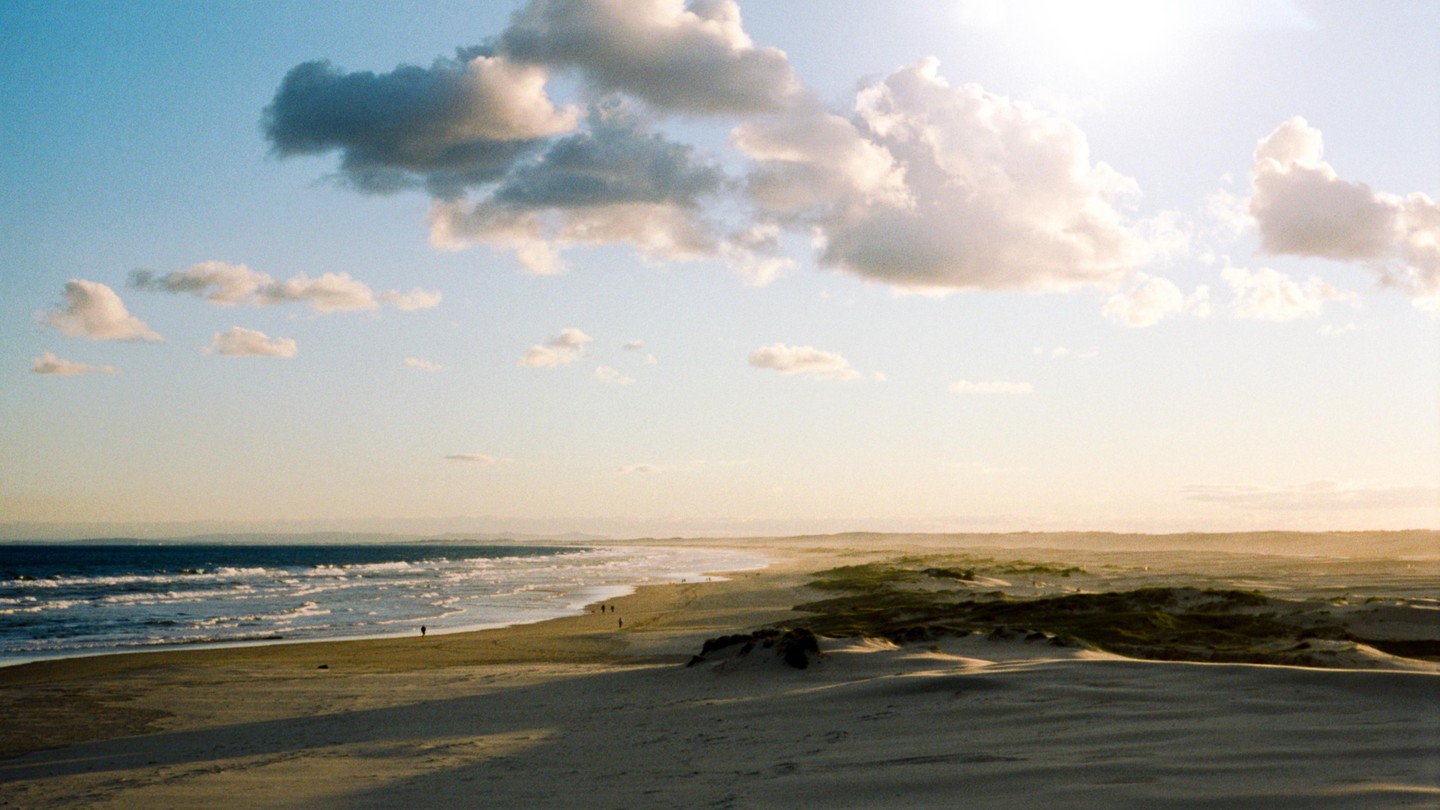 A wide view shows a beach with waves crashing on the shore, sand dunes, and a bright sky with clouds.