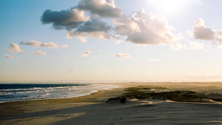 A wide view shows a beach with waves crashing on the shore, sand dunes, and a bright sky with clouds.