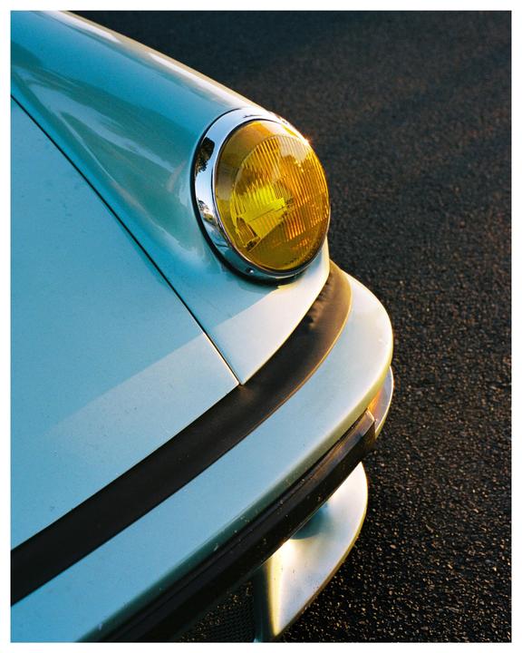 The front of a light blue car with a yellow headlight is shown from an elevated perspective.