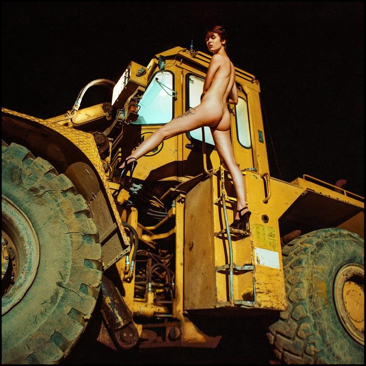 A nude woman in high heels stands on a ladder of a large yellow construction vehicle at night.