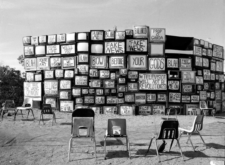 A wall made of stacked televisions with various phrases written on their screens stands behind several empty chairs on a dirt ground.