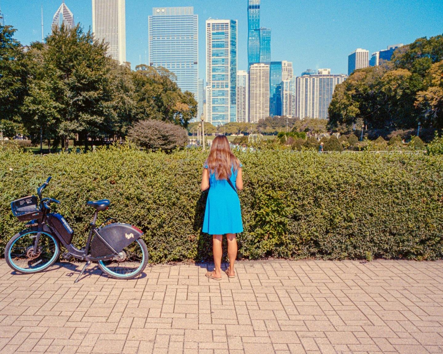 A woman in a blue dress stands on a paved path next to a bicycle and a hedge, looking towards a city skyline in the distance.