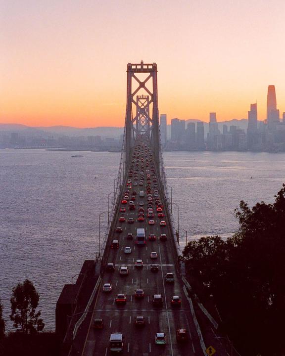 A bridge full of cars leads to a city skyline at sunset.