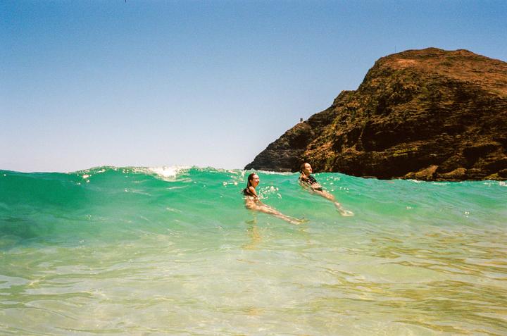 Two women are swimming in clear turquoise water with a large rocky hill in the background.