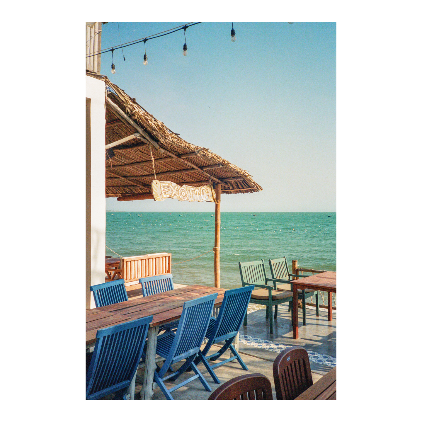 A thatched roof covers a patio with tables and chairs overlooking the ocean.