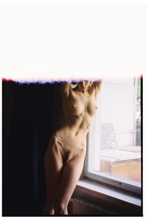 A nude woman stands with her arms raised, leaning against a wall next to a window.
