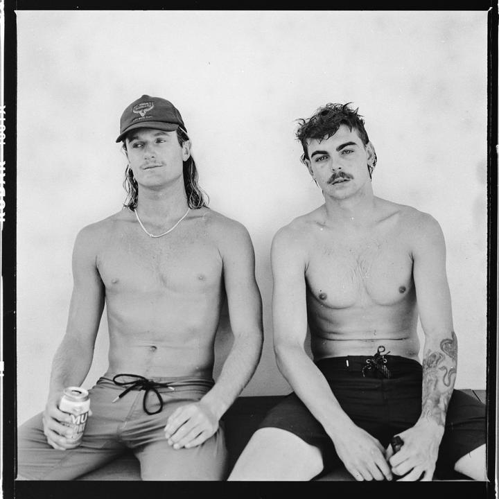 Two men sit shirtless, one wearing a cap and holding a can, the other with a mustache and a tattoo on his arm.