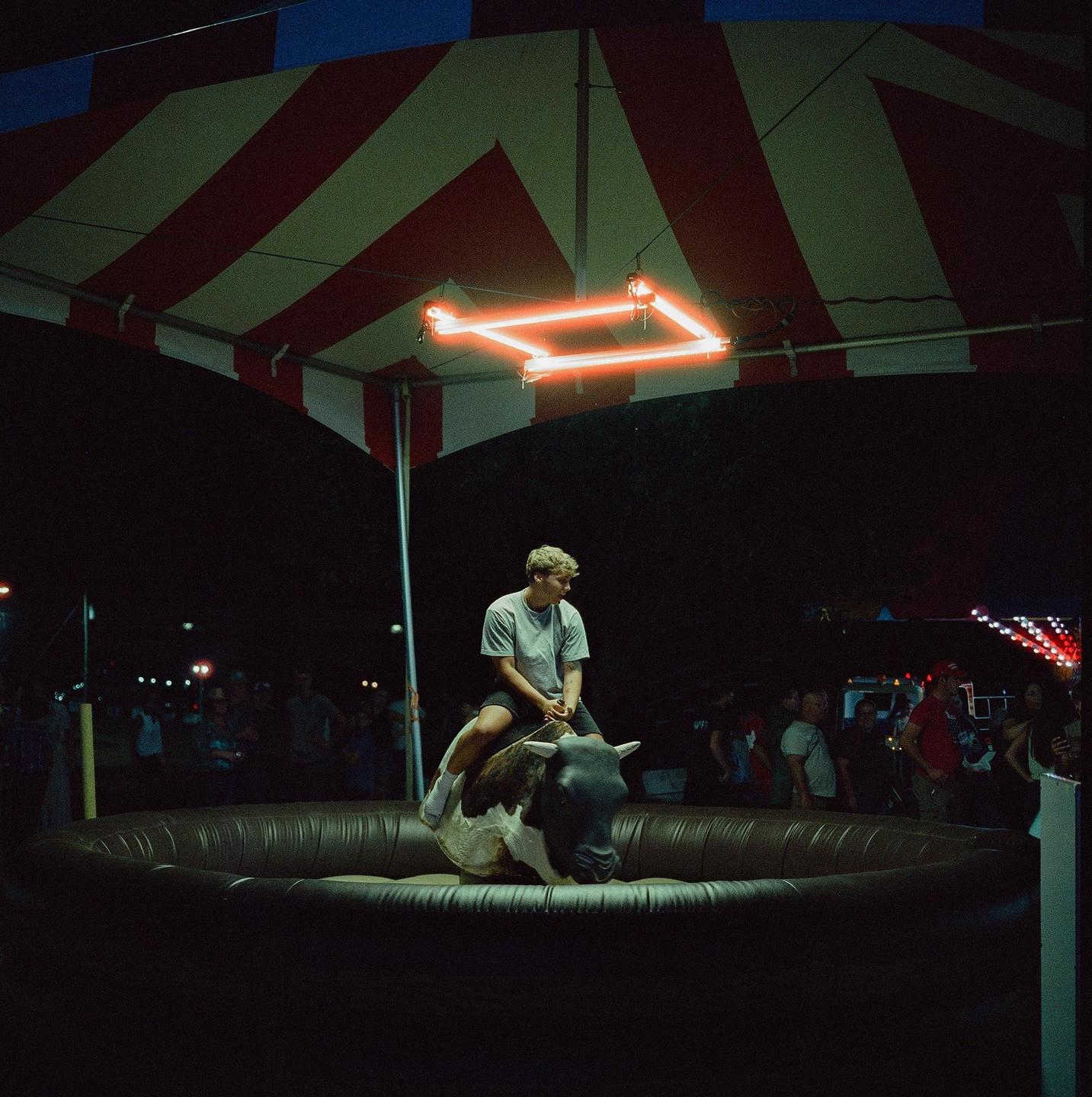 A man rides a mechanical bull under a striped tent with neon lights at night.