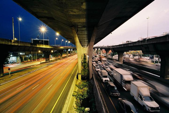 A highway overpass at night shows light trails from moving vehicles on the left and blurred traffic on the right.