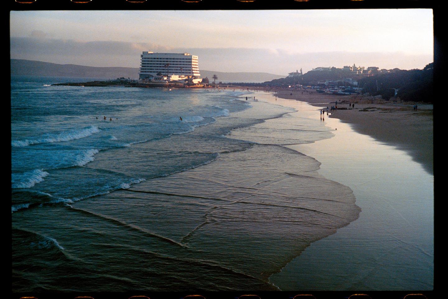 Waves roll onto a beach with a large hotel and a town on a hill in the background at sunset.