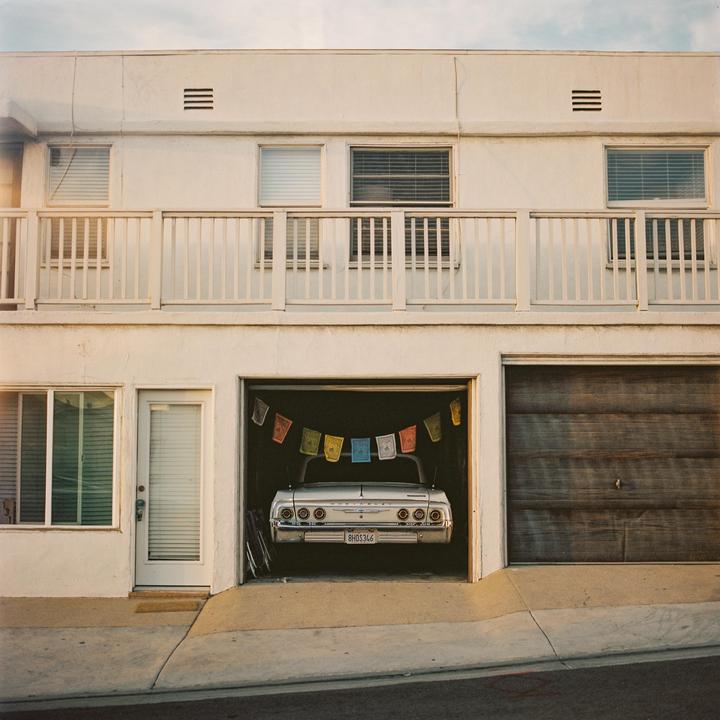 A white car is parked in a garage with prayer flags hanging above it, next to a closed wooden garage door and a white door.