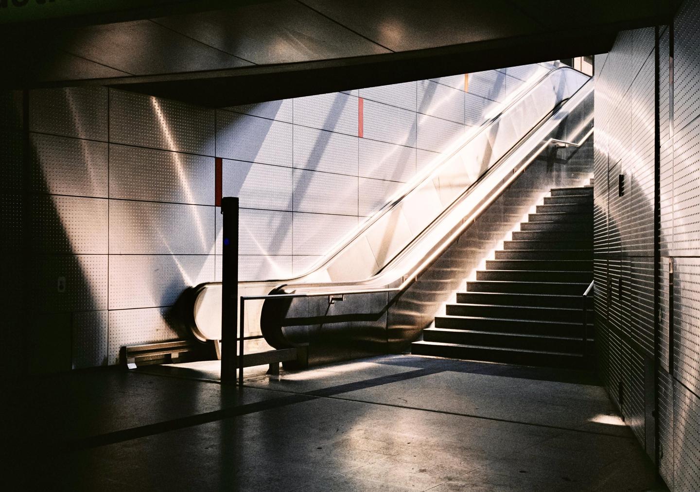 Sunlight casts strong shadows on an escalator and staircase in a subway station.