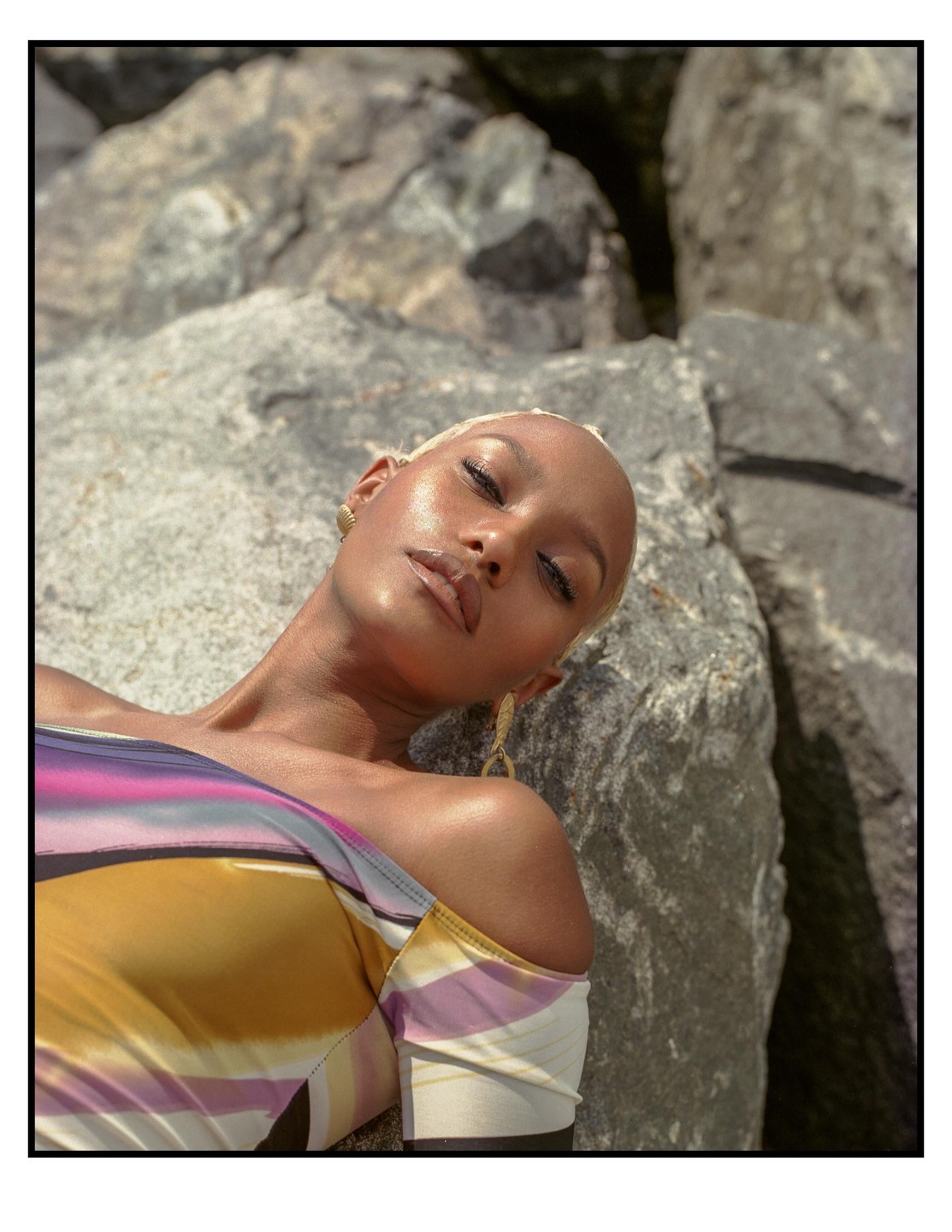 A woman with short blonde hair and a colorful top is lying on rocks.