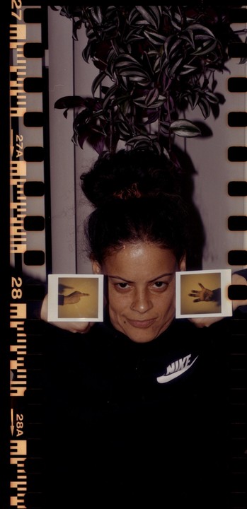 A woman holds two instant photos of hands in front of her face, with a plant visible above her head.