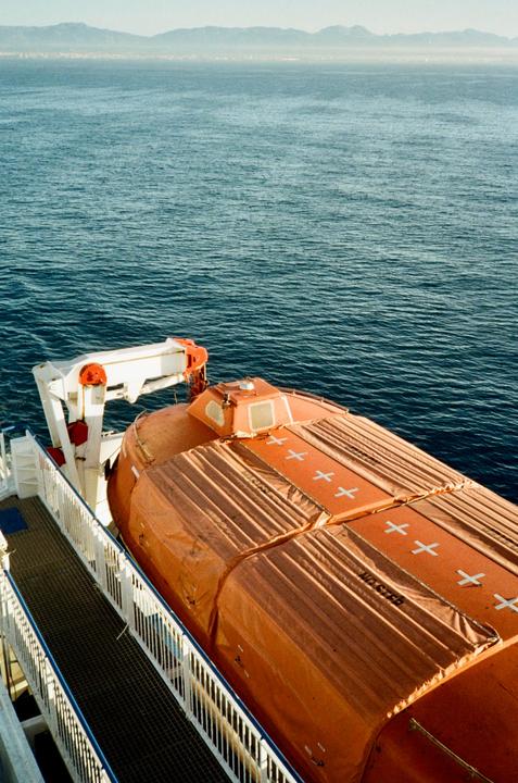 An orange lifeboat is visible on the side of a ferry, with the sea and distant mountains in the background.