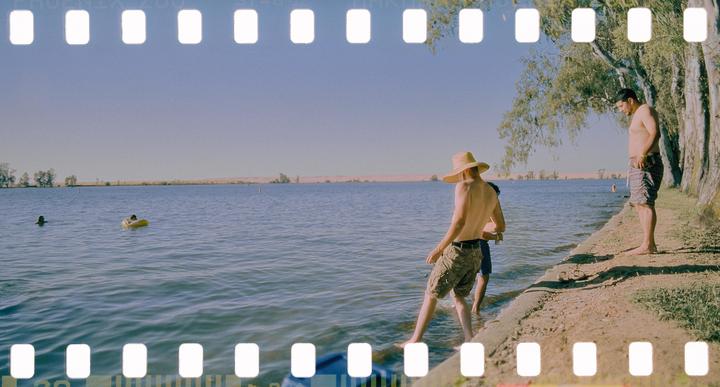 Three men are by a lake, with two in the water and one standing on the shore.