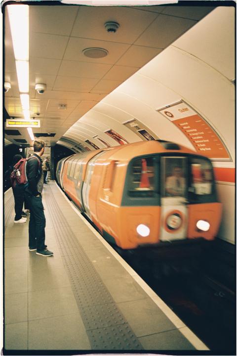 A man with a backpack stands on a subway platform as an orange train approaches.