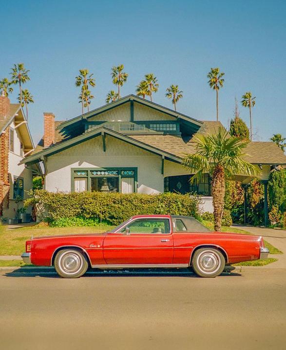 A red car is parked on a street in front of a house with palm trees in the background.
