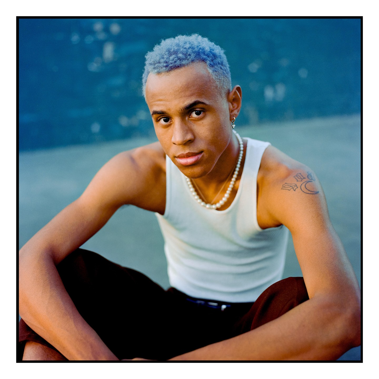 A man with blue hair and a pearl necklace sits looking at the viewer.