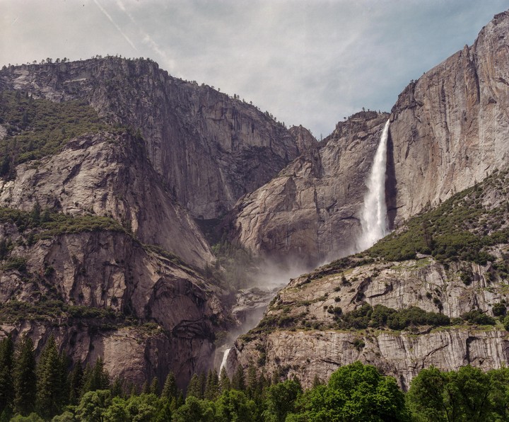 A tall waterfall cascades down a rocky mountain face, surrounded by green trees.