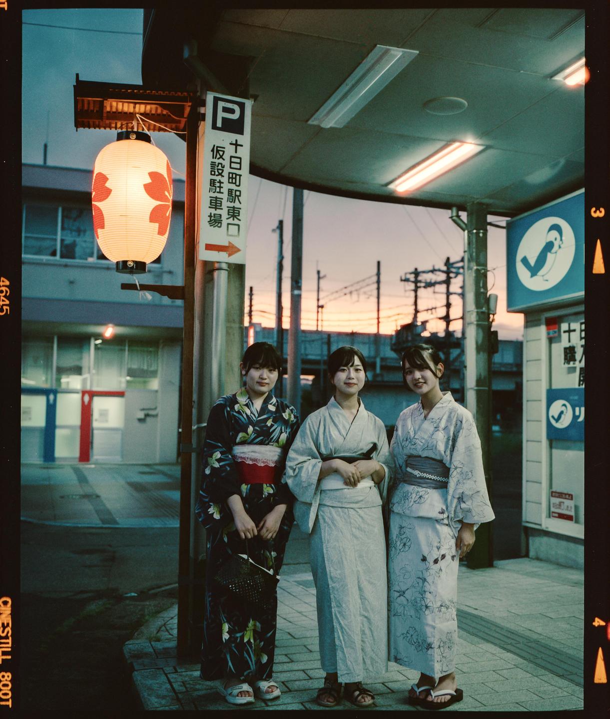 Three women in traditional Japanese clothing stand on a street at dusk next to a paper lantern.