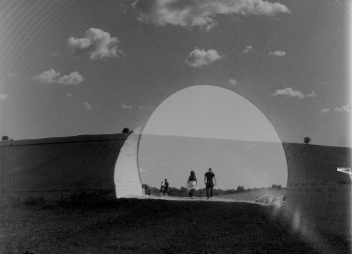 Three people walk on a path next to a large curved structure under a sky with clouds.