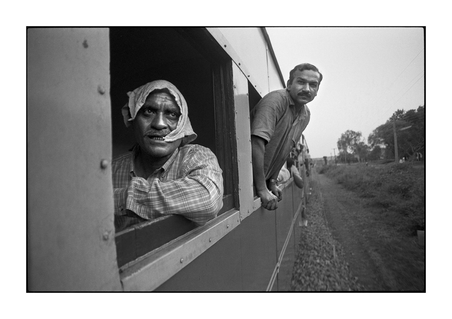 Two men look out of the windows of a moving train, with one man wearing a head covering and the other leaning out.