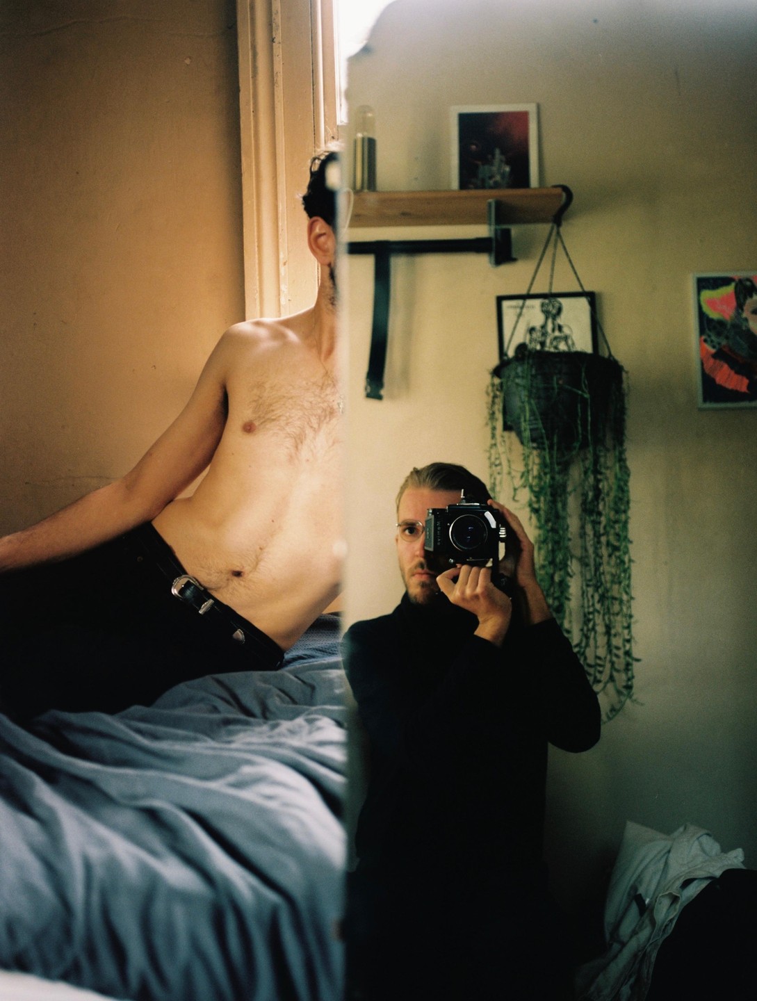 A man with a bare torso is reflected in a mirror, while another man in a black turtleneck takes a self-portrait with a camera.