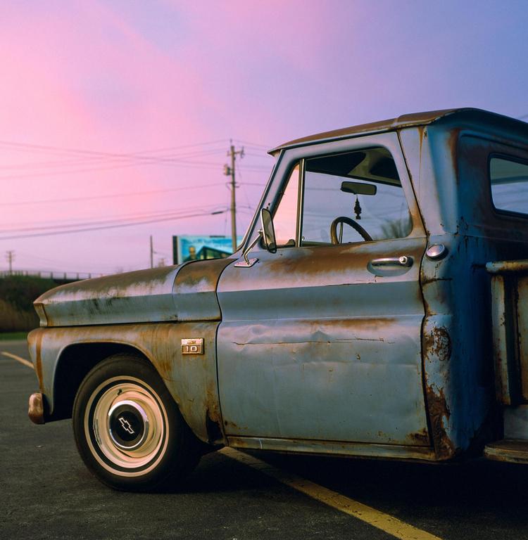 A rusty blue pickup truck is parked on asphalt with a pink and purple sky in the background.